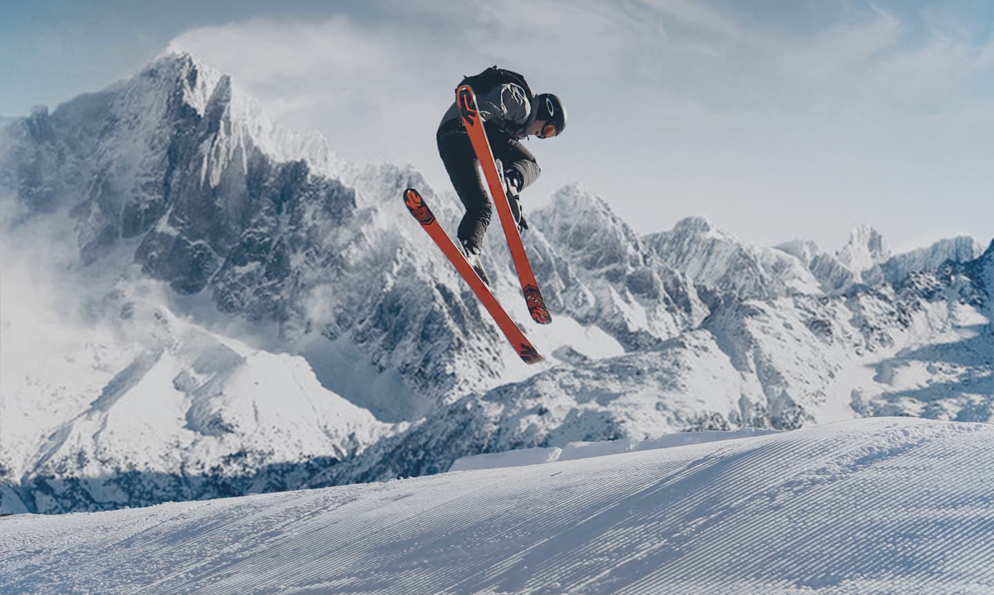 Skier catching big air mid-jump against a snow-capped mountain range.