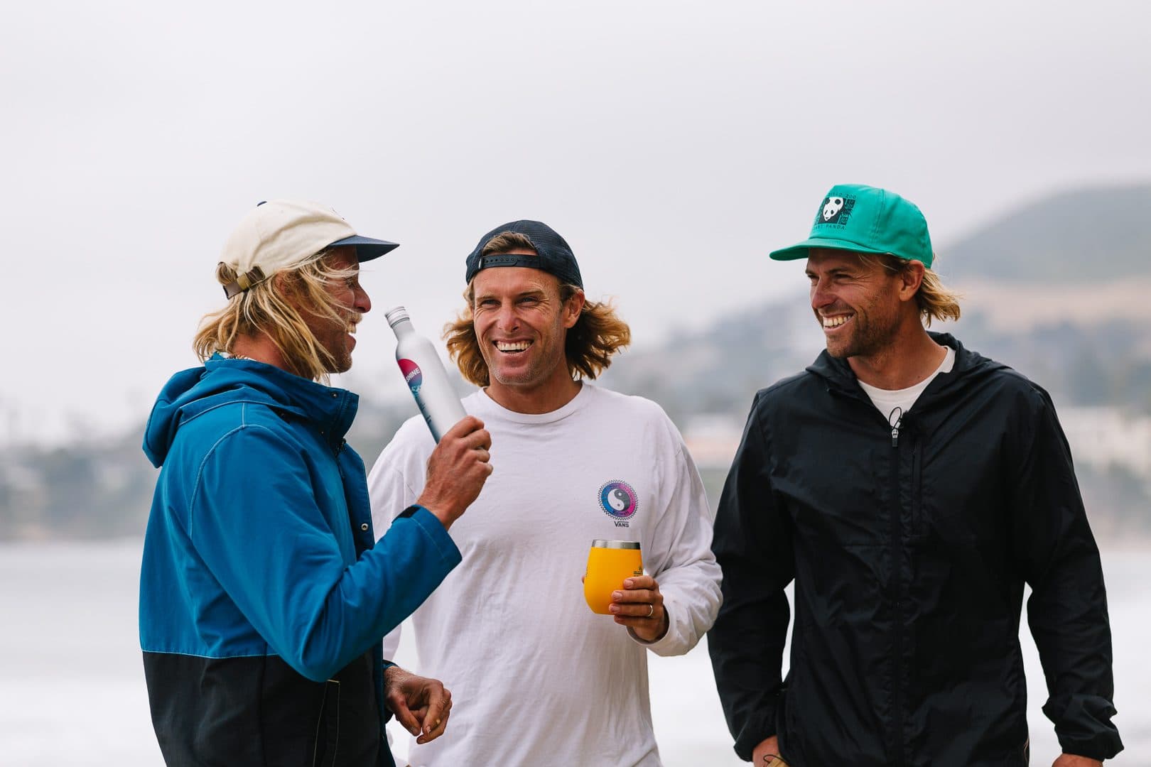 Three men laughing together outdoors, one holding a canned beverage and an insulated tumbler