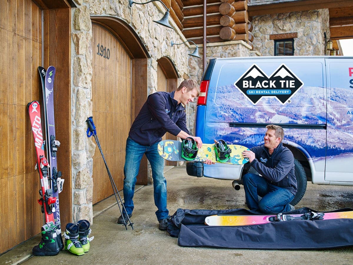 Black Tie Ski Rental Delivery staff loading snowboard into van beside skis