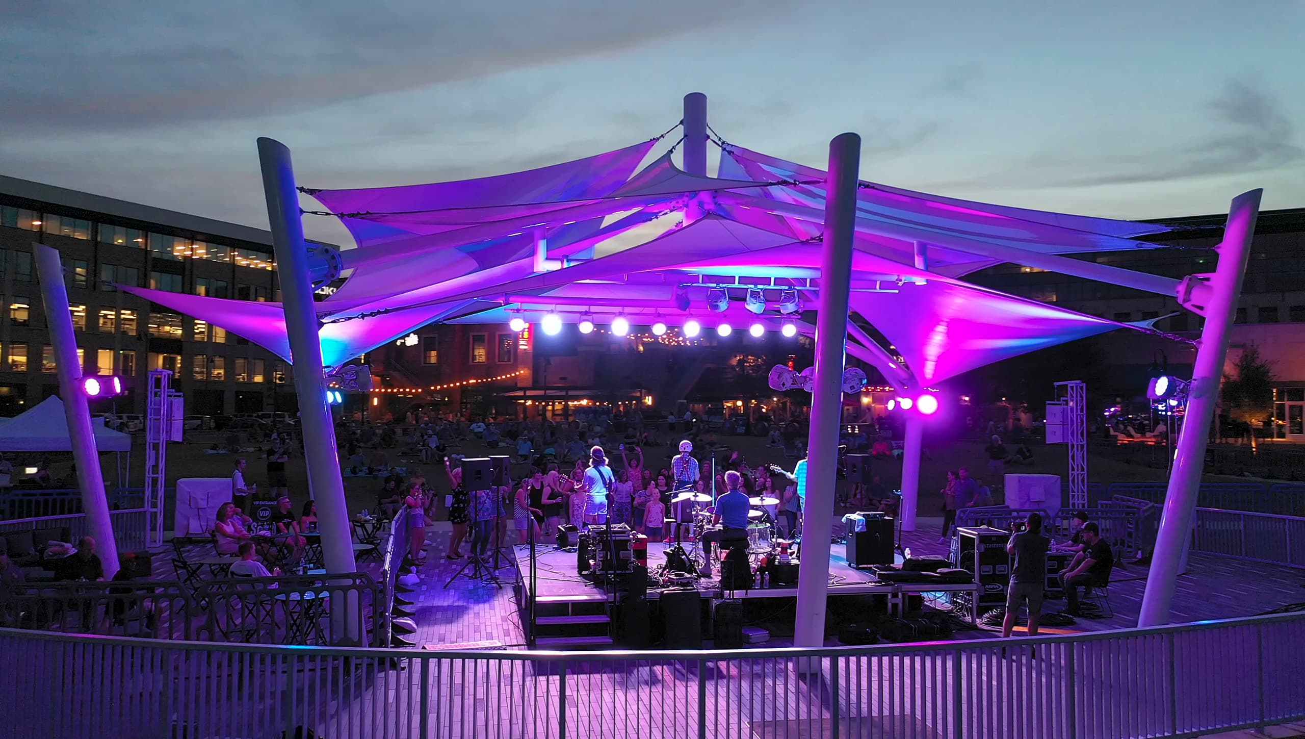 Purple-lit fabric shade canopy over a live band performing at an outdoor night concert
