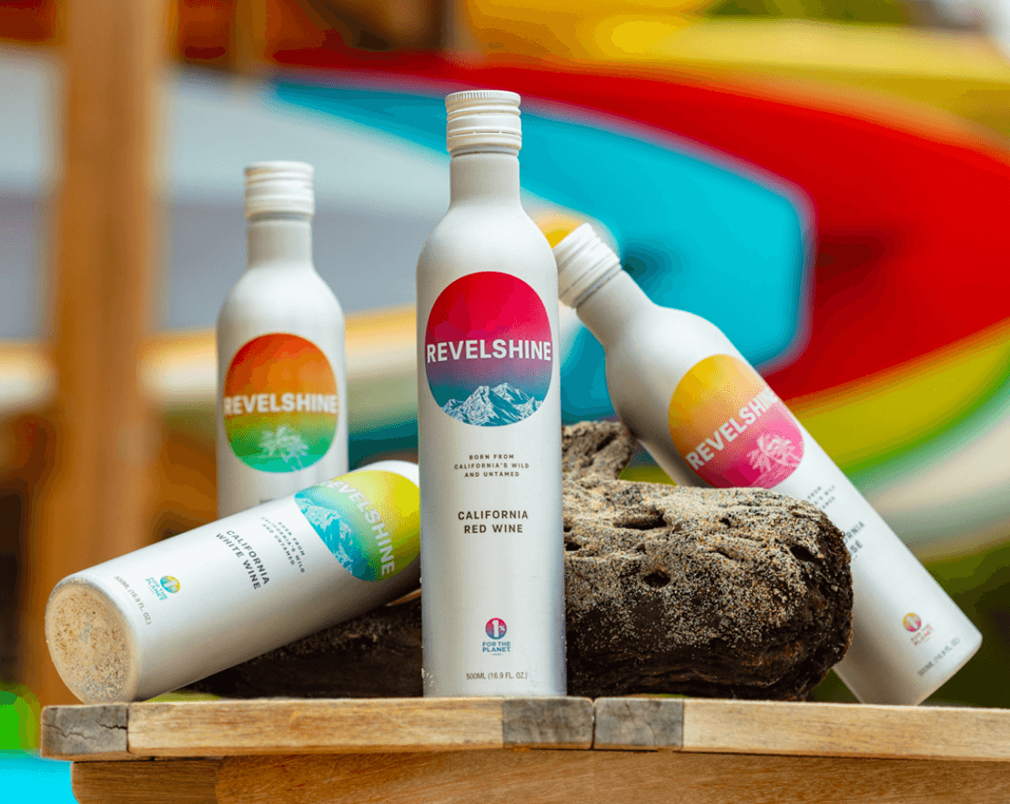 Four Revelshine wine bottles arranged on driftwood with colorful kayaks in the background