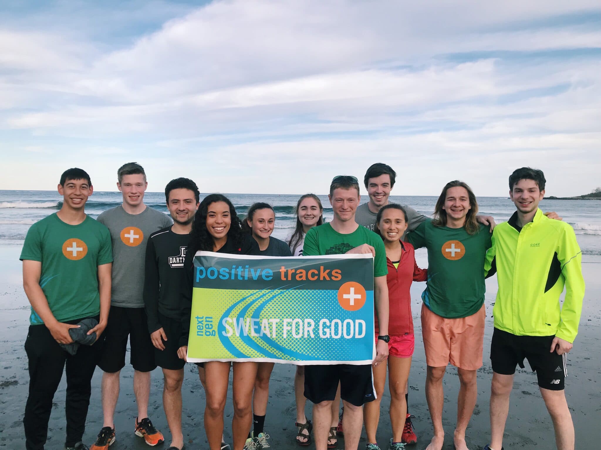 Group of young volunteers on a beach holding a 'Positive Tracks Sweat For Good' banner