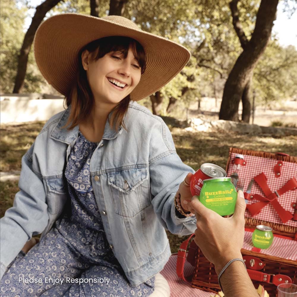 Woman in a sunhat clinking BuzzBallz cocktail cans during an outdoor picnic
